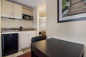 A compact kitchen with a refrigerator, microwave, and a coffee maker. There is a dining table with chairs and a framed picture on the wall.