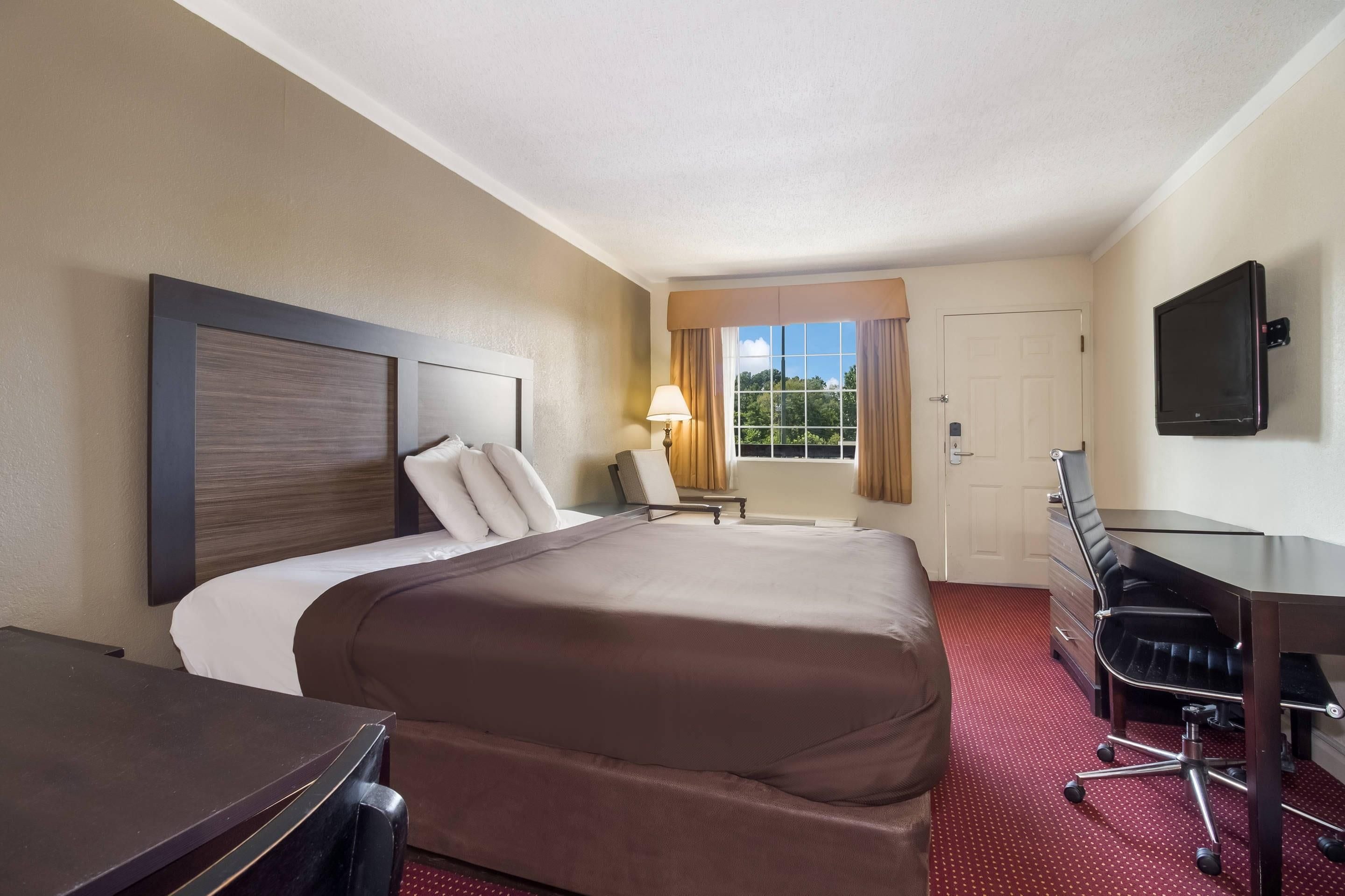 Ein Hotelzimmer mit einem großen Bett, einem Schreibtisch, einem Sessel, einem Fernseher und einem Fenster mit Vorhängen.