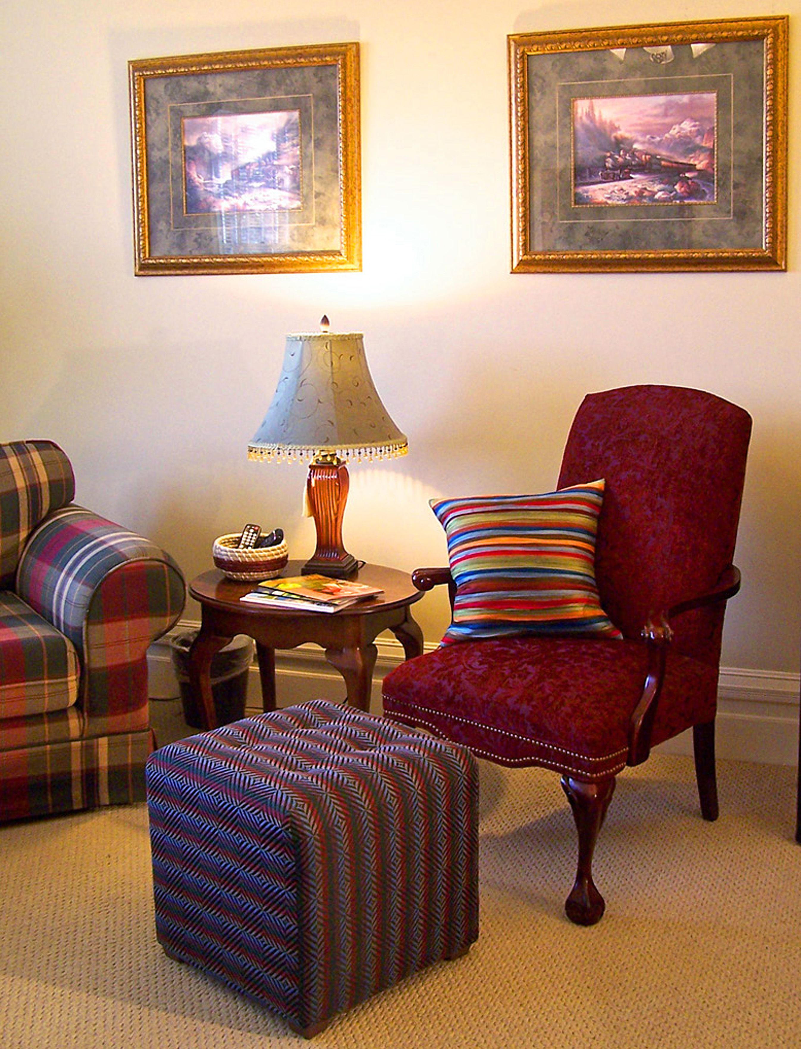 A living room with a plaid sofa, a red armchair with a striped pillow, a patterned ottoman, a table with a lamp, and two framed pictures on the wall.