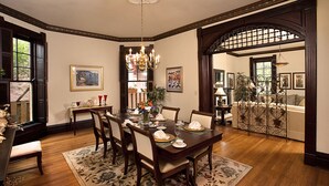 A dining room with a wooden table set for four, surrounded by chairs. There is a chandelier above the table and a large window to the left.