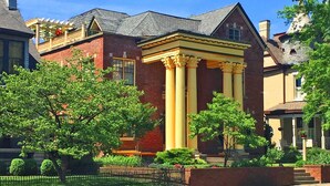 A brick house with a yellow porch and columns, surrounded by greenery and a fenced yard.