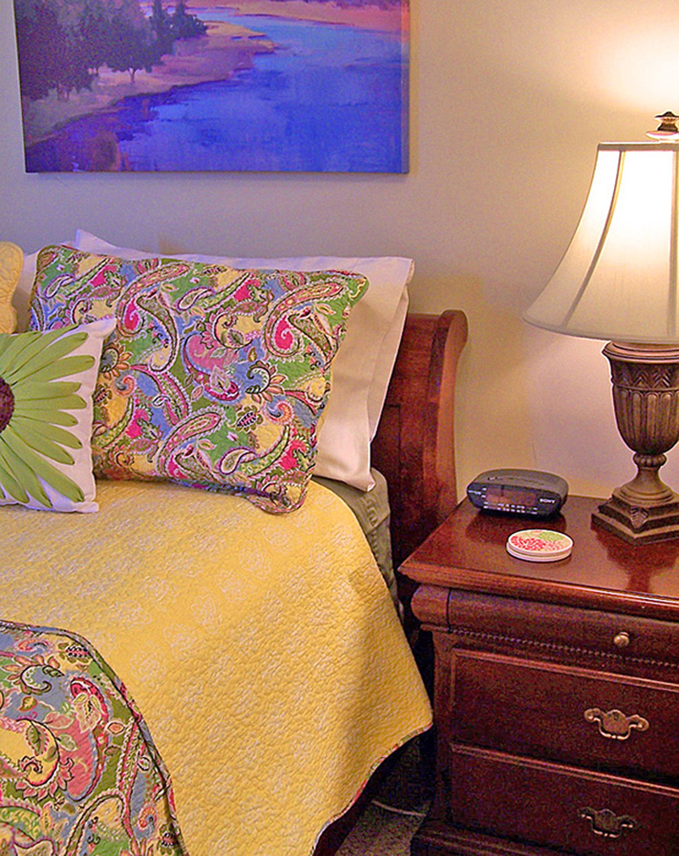 Una habitación de hotel con una cama, almohadas coloridas, una mesita de noche con una lámpara y un cuadro en la pared.