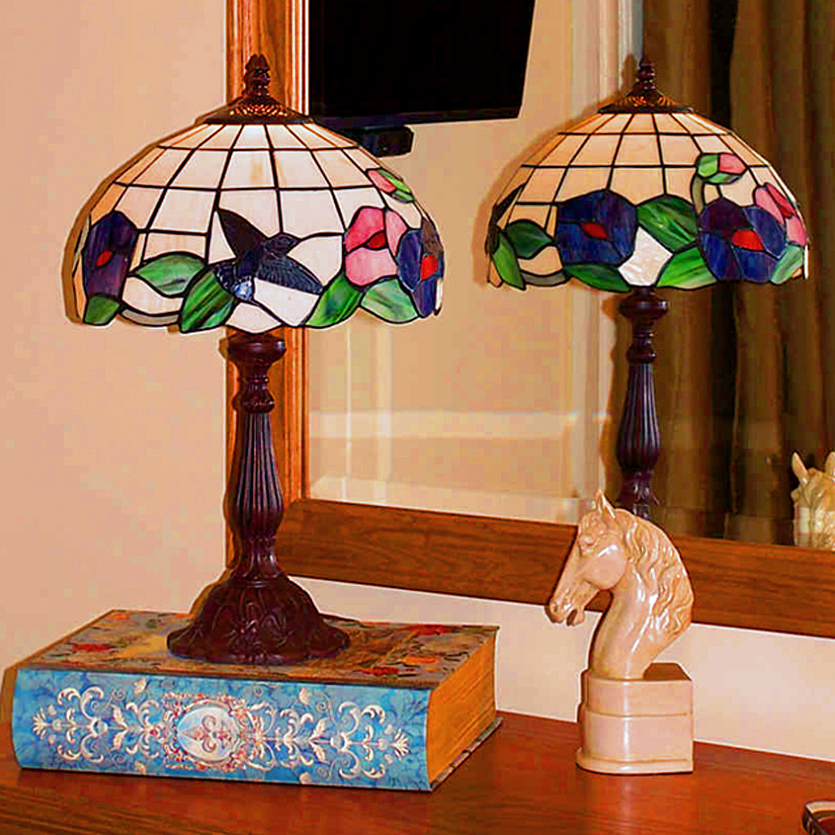 Duas luminárias de vitral com motivos florais sobre uma mesa de madeira, ao lado de um livro e uma estatueta de cavalo.