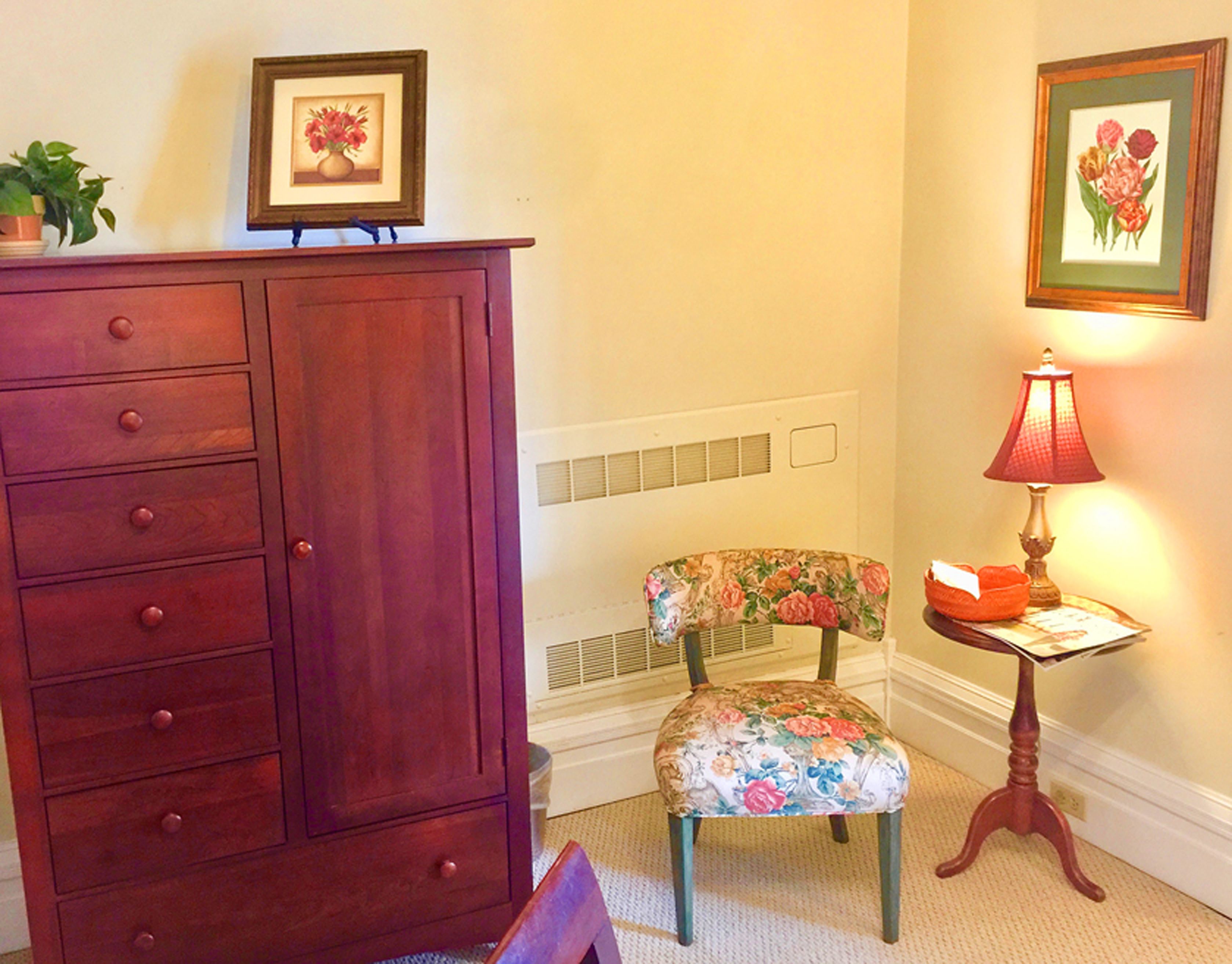 Una habitación con un armario de madera, un sillón floral, una mesa con lámpara y cuadros en la pared.