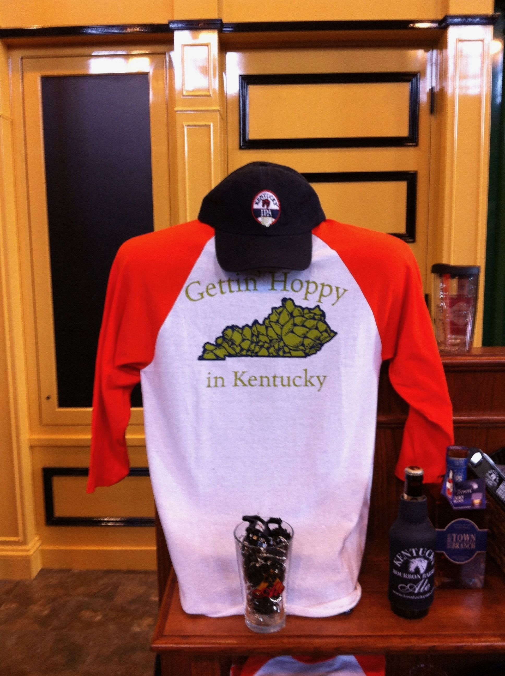 A T-shirt with "Gettin' Hoppy in Kentucky" printed on it, placed on a table with a bottle of Kentucky Bourbon and a glass of beer.