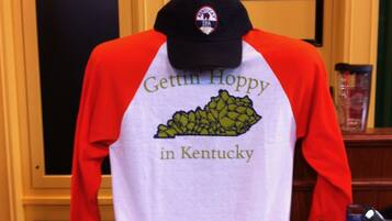 A T-shirt with "Gettin' Hoppy in Kentucky" printed on it, placed on a table with a bottle of Kentucky Bourbon and a glass of beer.