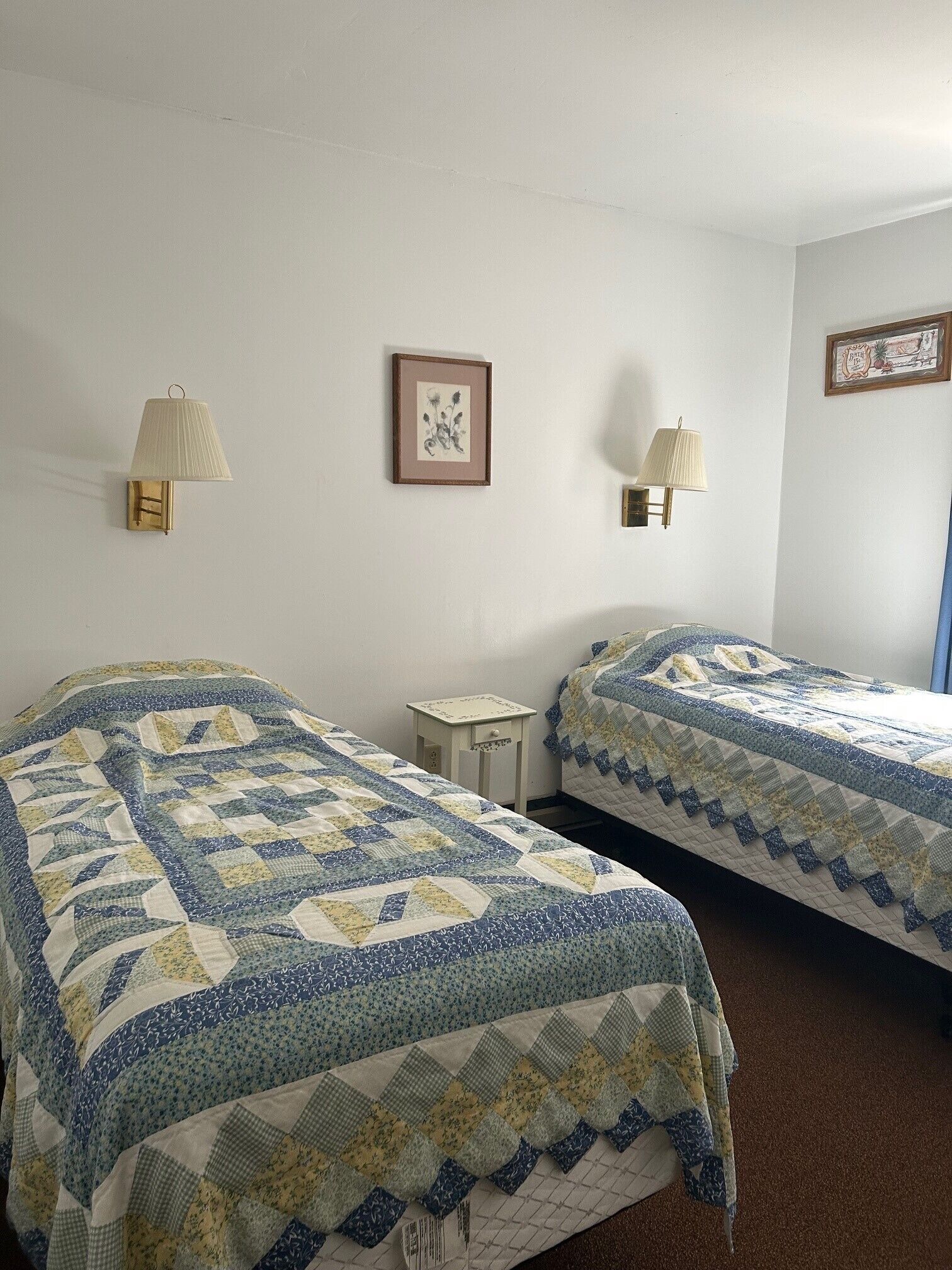 Una habitación con dos camas, cada una con colcha azul y blanca, una mesita de noche pequeña y una lámpara de pared.
