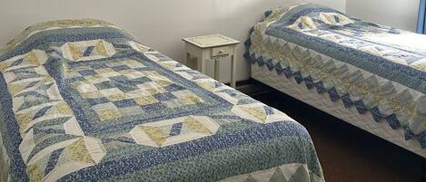 Una habitación con dos camas, cada una con colcha azul y blanca, una mesita de noche pequeña y una lámpara de pared.