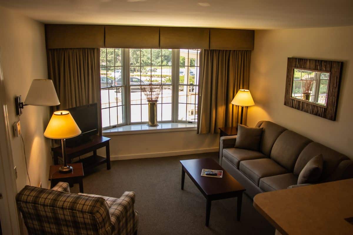 Quarto de hotel com sofá, poltrona, mesa de centro, televisão e luminárias.