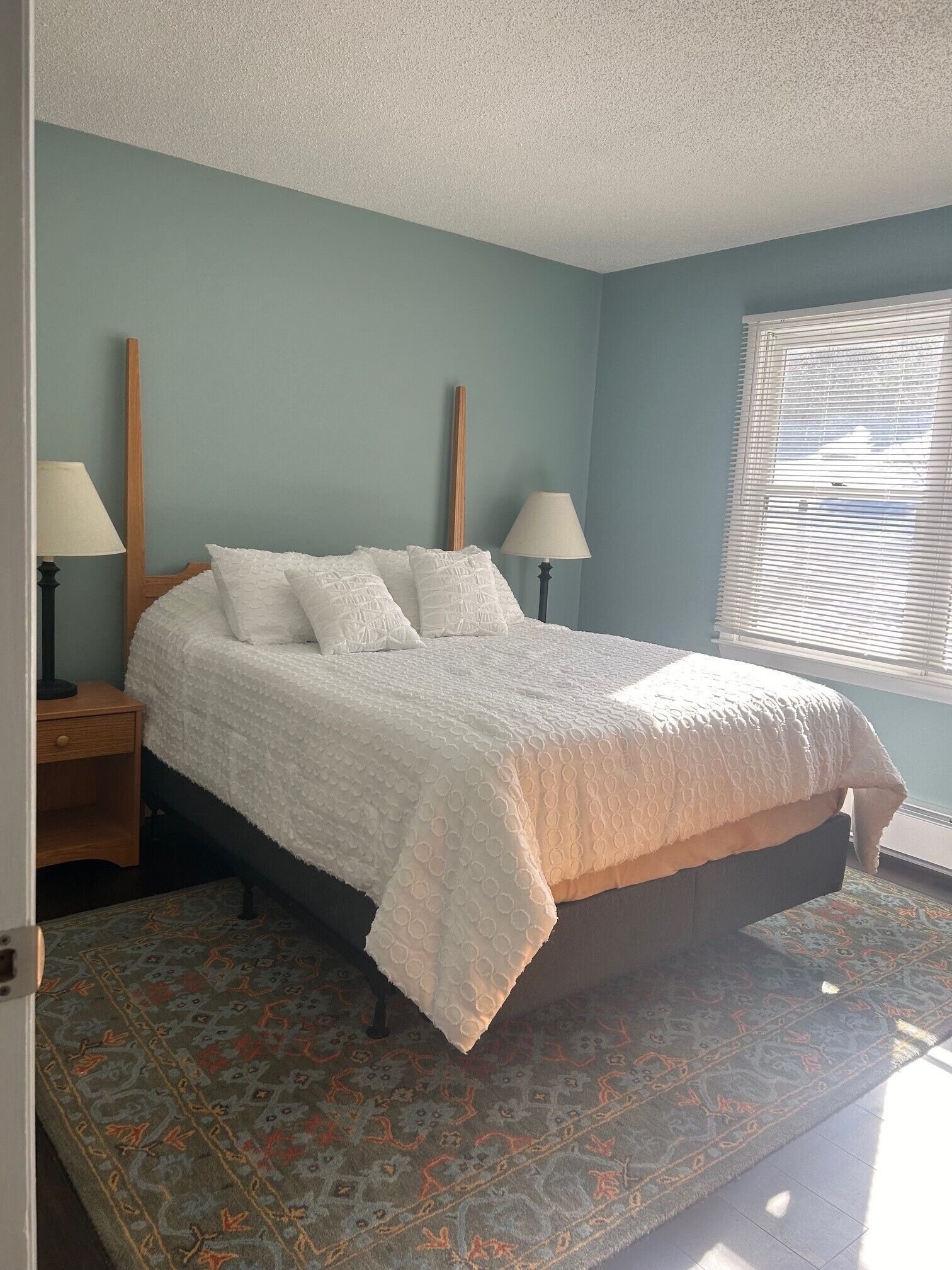 A bedroom with a bed, two bedside tables, a window with blinds, and a patterned rug.