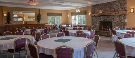 A spacious room with multiple round tables and purple chairs, a stone fireplace, and large windows.