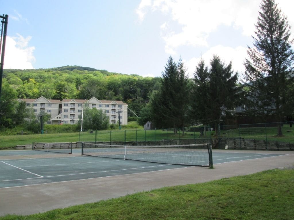 An outdoor tennis court with a net, surrounded by a grassy area and trees.