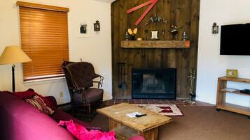 Un salon chaleureux avec une cheminée, un mur en bois, un fauteuil rouge, un canapé rose et un téléviseur.