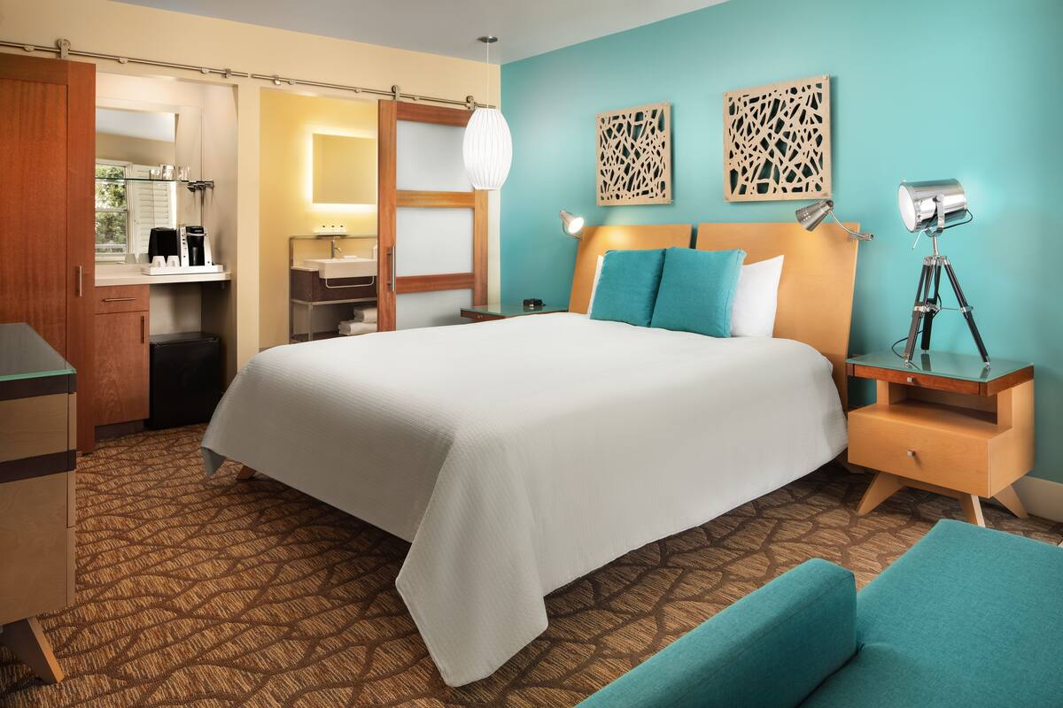 A hotel room with a large bed, a desk, a chair, a nightstand, a lamp, and a wall-mounted light fixture.