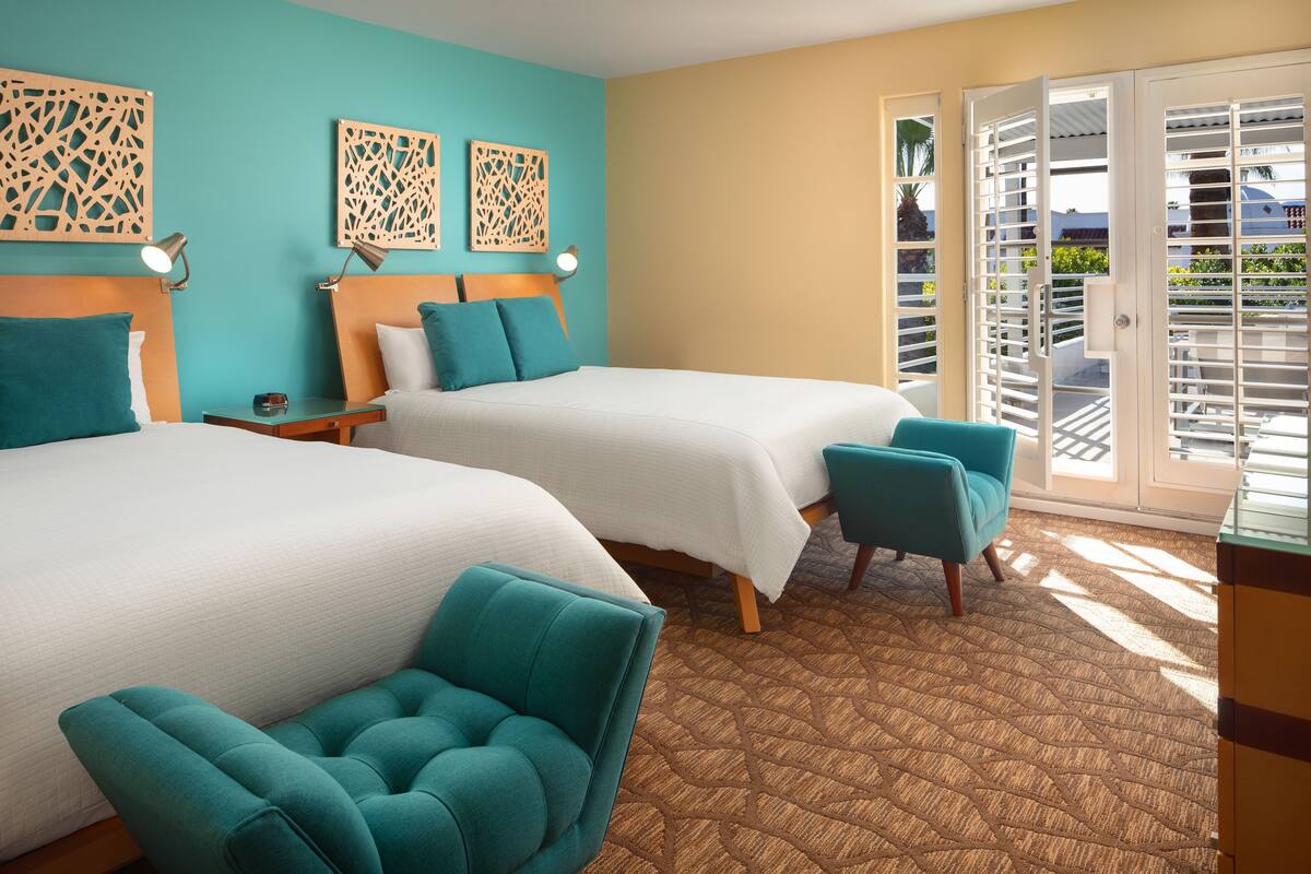 A hotel room with two beds, teal chairs, a wooden headboard, and a patterned carpet.