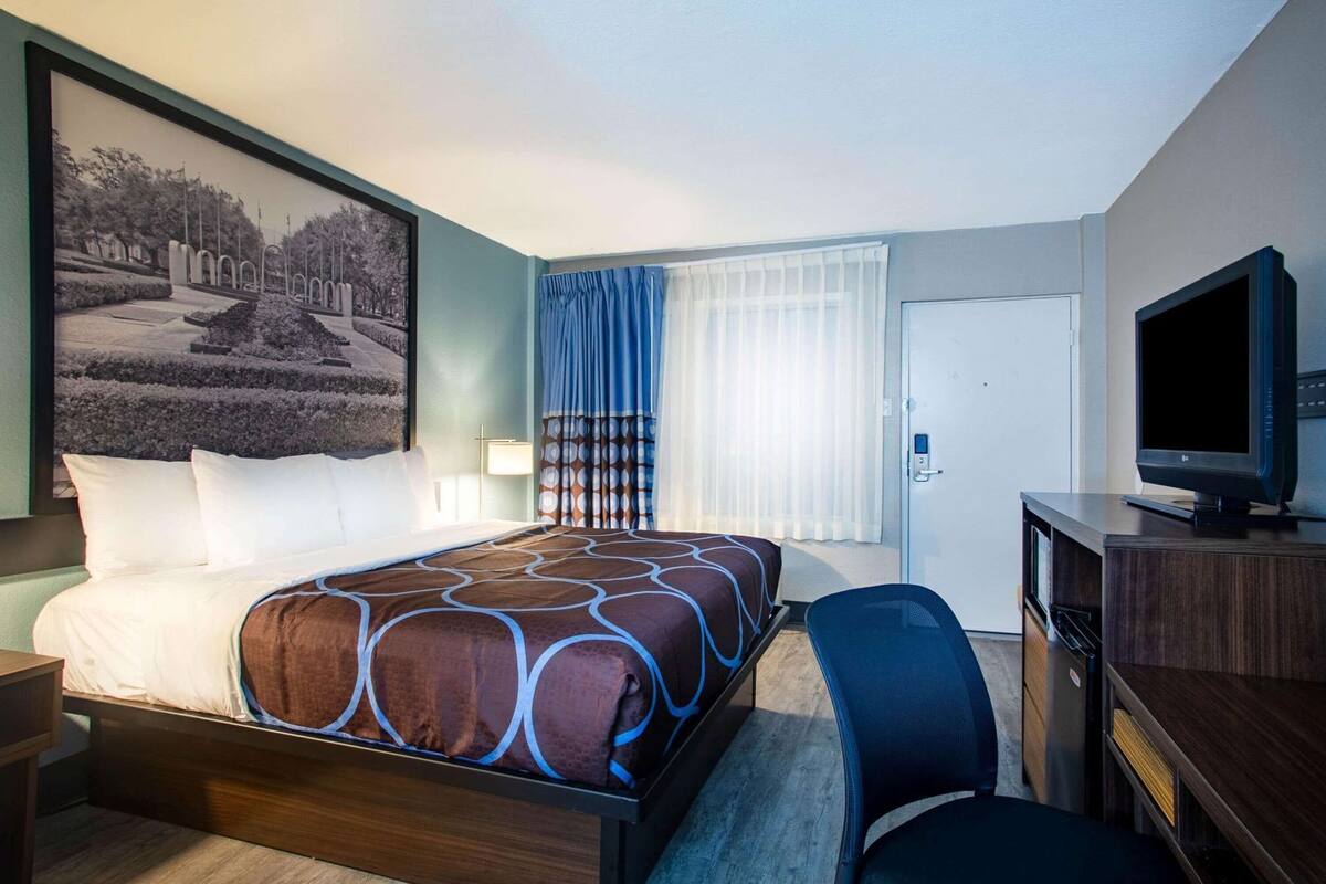 A hotel room with a bed, a desk with a television, a chair, a door, and a window with curtains.
