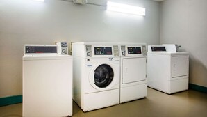 A laundry area with four washing machines and dryers, fluorescent lighting, and a plain wall. - Super 8 by Wyndham Mobile I-65 (Mobile)