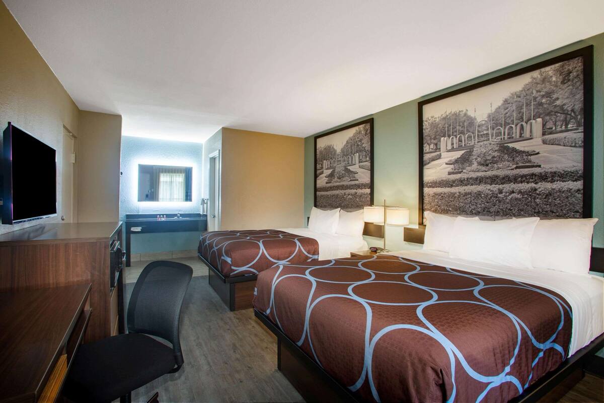 A hotel room with two beds, a desk, a chair, a TV, and large framed pictures on the wall.
