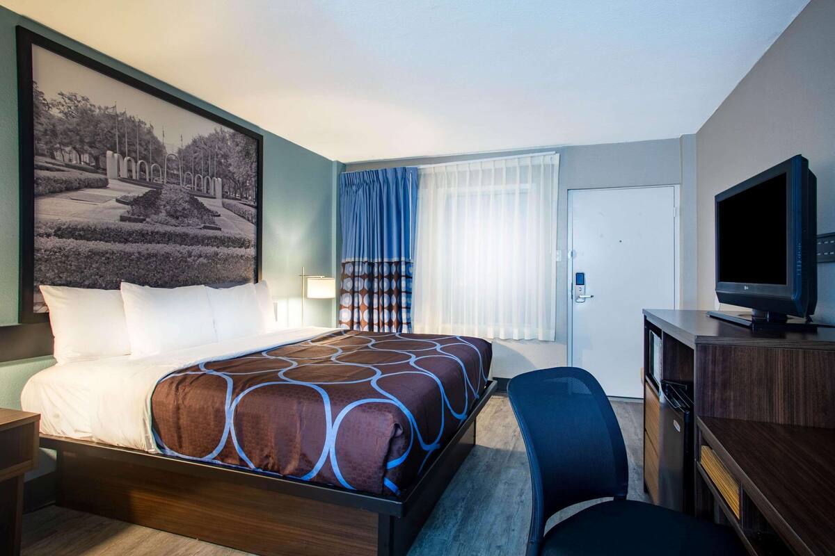 A hotel room with a large bed, a desk with a television, a chair, and a window with curtains.