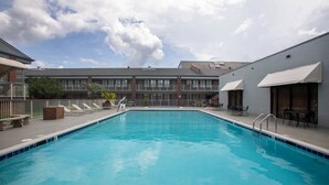 A rectangular swimming pool with a tiled edge, surrounded by lounge chairs and tables, with a building featuring large windows and awnings in the background. - Super 8 by Wyndham Mobile I-65 (Mobile)