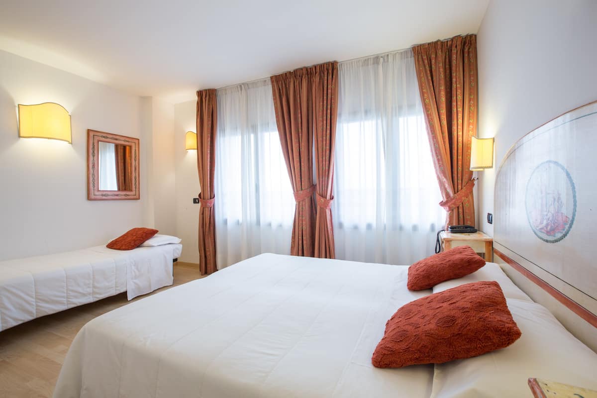 A hotel room with two single beds, a large bed, and a headboard with a circular design. There are curtains, a mirror, and wall-mounted lamps.