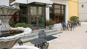 An outdoor seating area with tables and chairs, a decorative stone fountain, and a building entrance with glass doors and a white awning. - Hotel Ariston (Mestre)