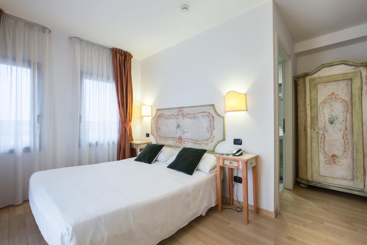 A neatly made bed with white linens and two green pillows, a bedside table with a lamp, and a wooden wardrobe with decorative paneling.