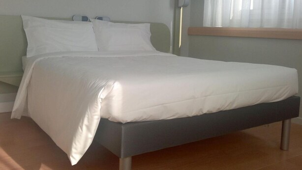Una cama bien hecha con ropa de cama blanca en una habitación con piso de madera y pared de color claro.