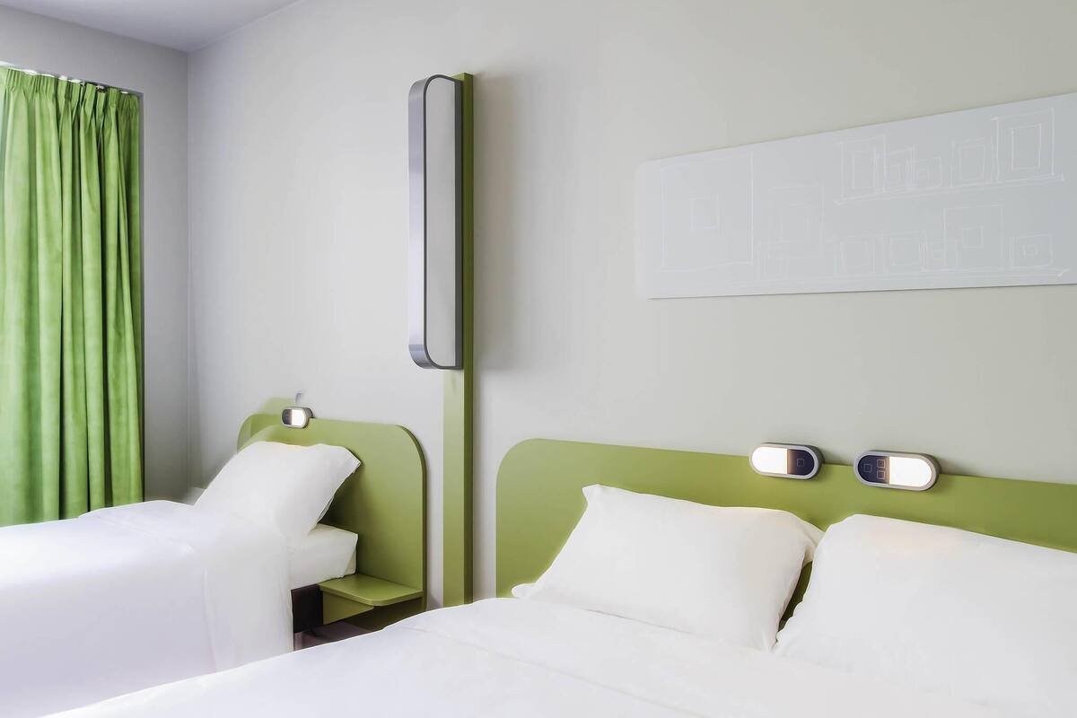 A hotel room with two beds, a green headboard, a bedside table with a lamp, and a wall-mounted light.