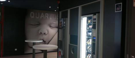 A vending machine with a variety of snacks and drinks, a small table, and a wall with a large advertisement.