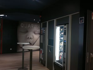 A vending machine with a variety of snacks and drinks, a small table, and a wall with a large advertisement. - ibis budget Braga Centro (Braga)