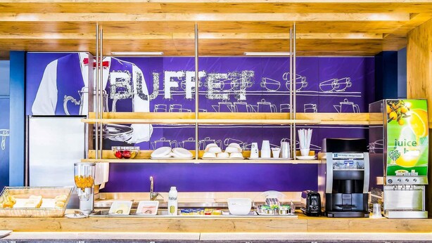 A buffet counter with a purple backdrop, various food items, a juice dispenser, and a coffee machine. - ibis budget Braga Centro (Braga)