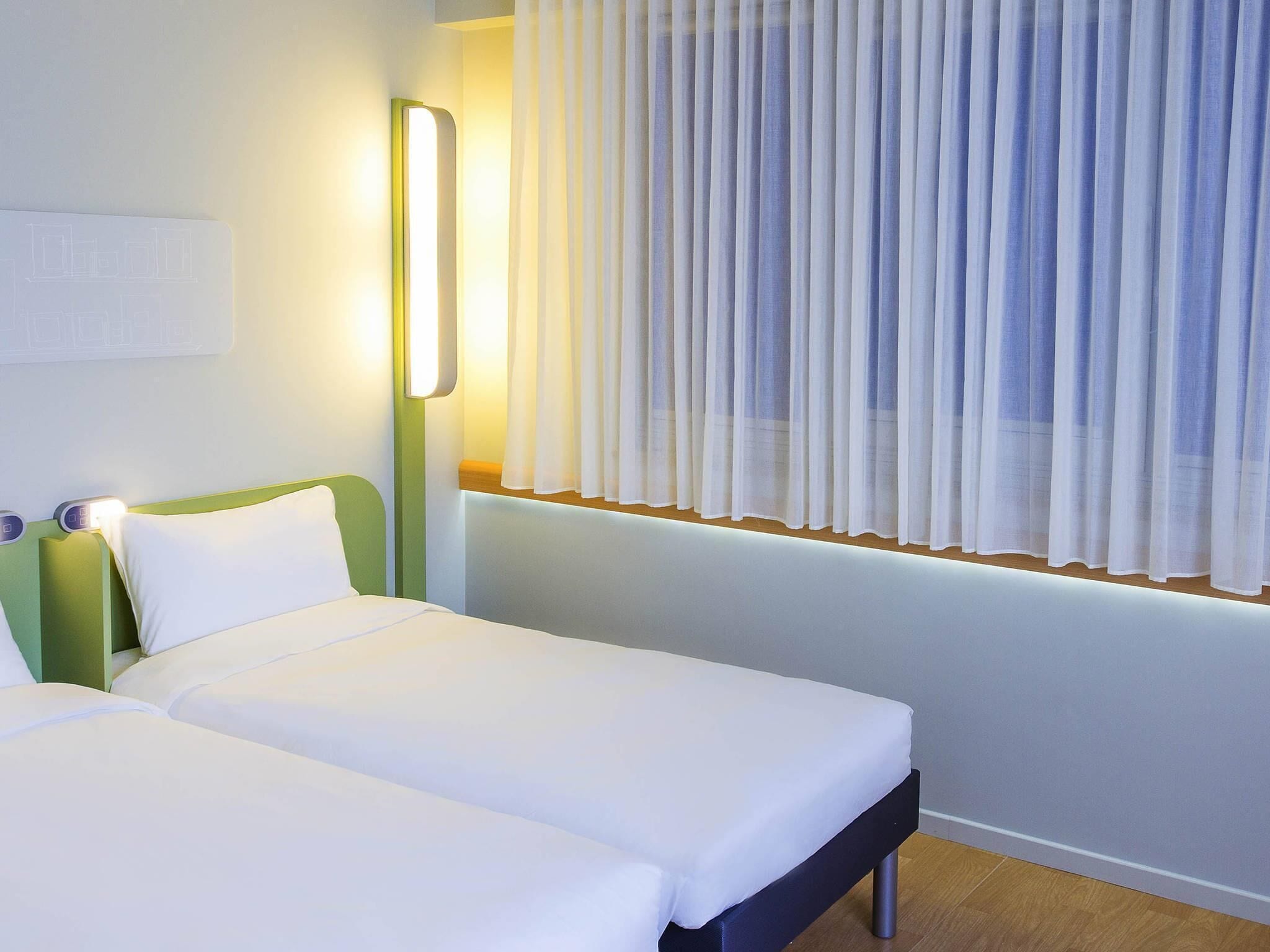 A hotel room with two beds, a bedside table, a mirror, and a window with sheer curtains.