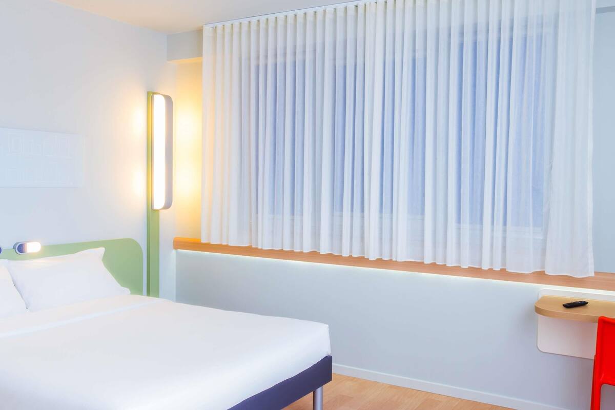 A hotel room with a bed, a bedside table, a chair, a window with curtains, and a wall-mounted light.