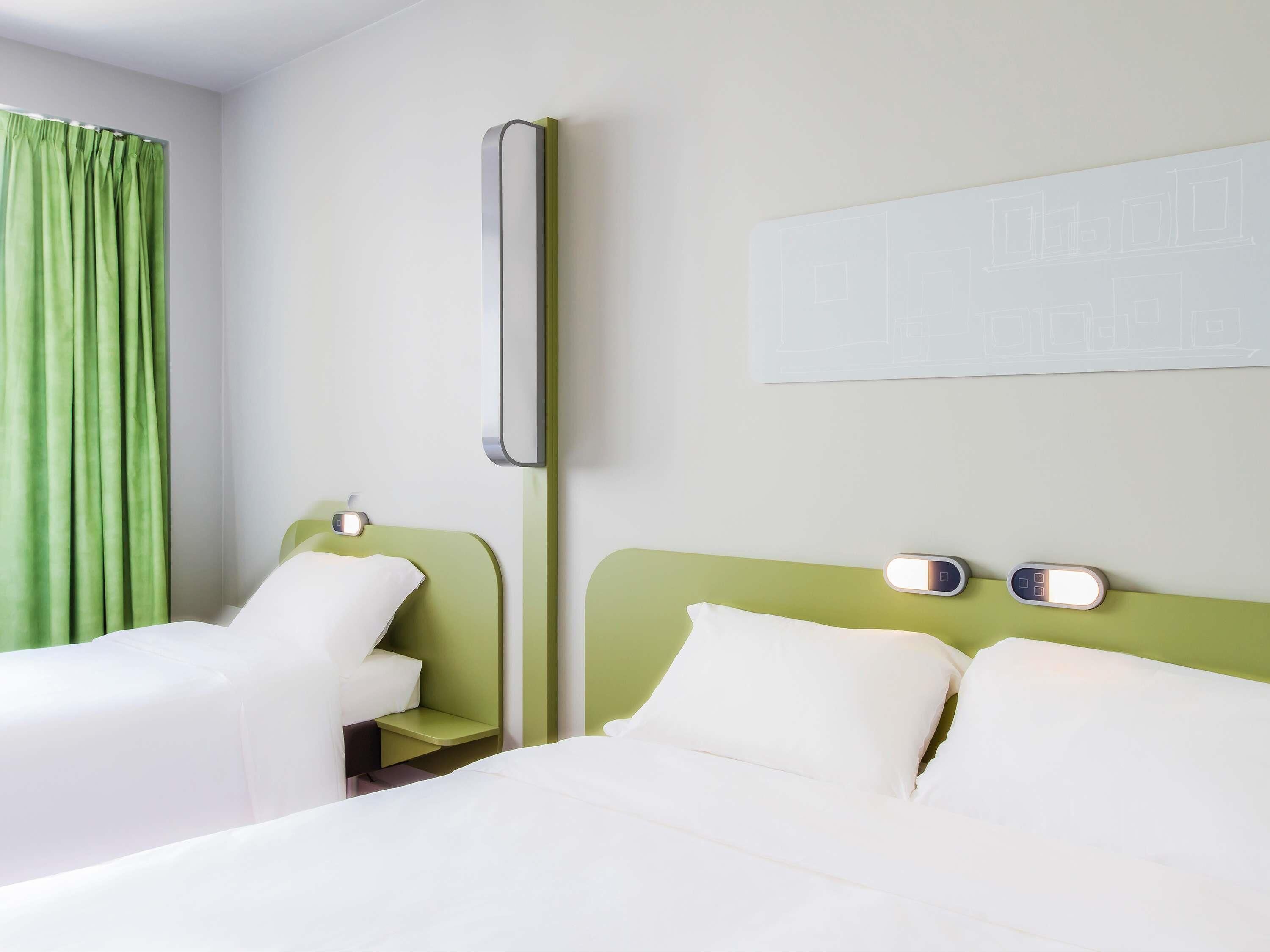 A hotel room with two beds, a green curtain, a wall-mounted light, and a white wall with a simple design.