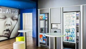 A modern interior with a blue wall, a large poster, vending machines, and a small table with a yellow stool. - ibis budget Braga Centro (Braga)