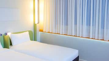 Habitación de hotel con dos camas, una mesita de noche con lámpara y una ventana con cortinas translúcidas.