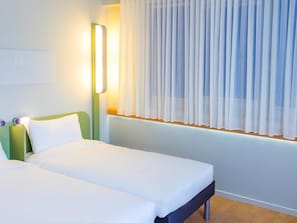 A hotel room with two beds, a bedside table with a lamp, and a window with sheer curtains. - ibis budget Braga Centro (Braga)