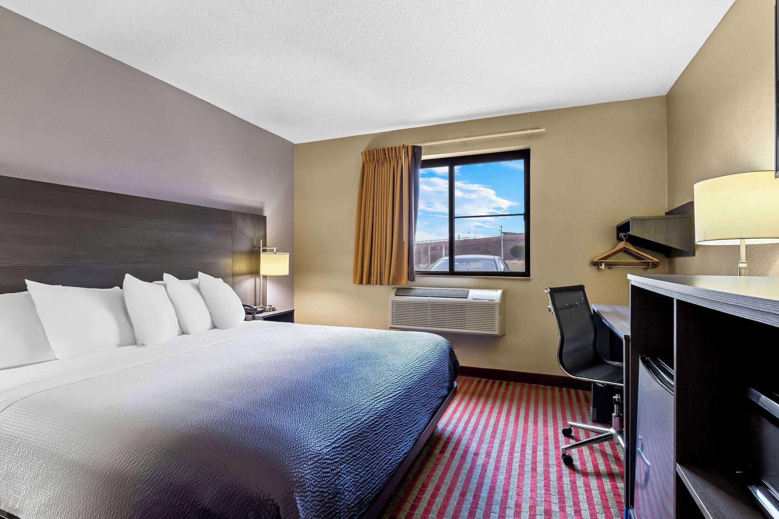 Habitación de hotel con una cama grande, un escritorio, una silla, un ventanal con cortinas y una lámpara.