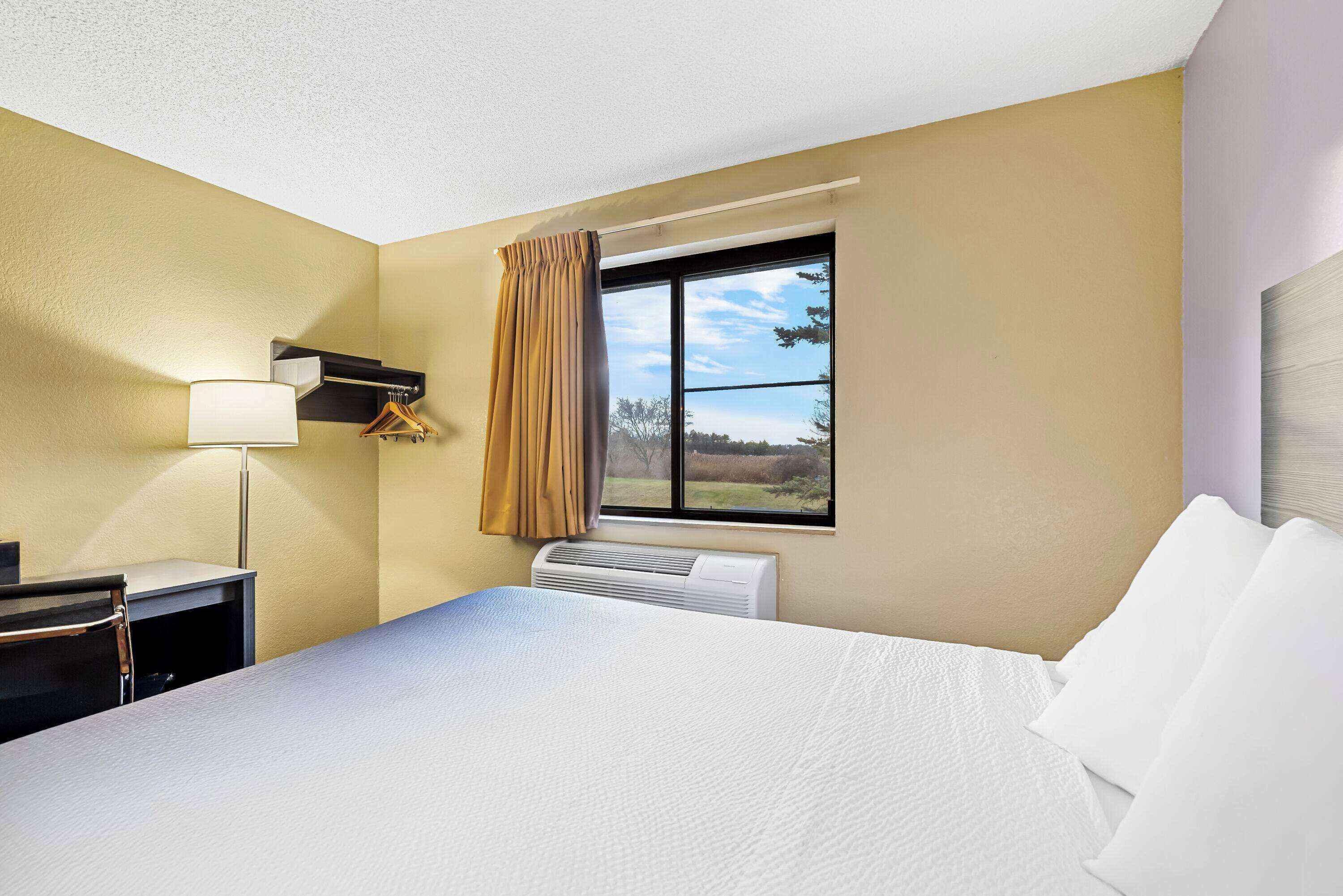 Habitación de hotel con cama, escritorio, lámpara, ventana con cortinas y equipo de aire acondicionado.