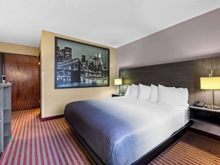 A hotel room with a large bed, two bedside lamps, a nightstand, a cityscape painting, and a wooden wardrobe.