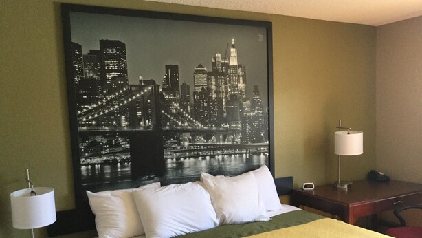 A hotel room with a bed, two bedside tables, a nightstand, a cityscape painting on the wall, and a desk with a lamp.