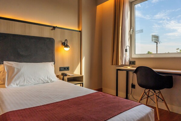 Habitación de hotel con cama, escritorio, silla, ventana con vistas y lámpara de pared.