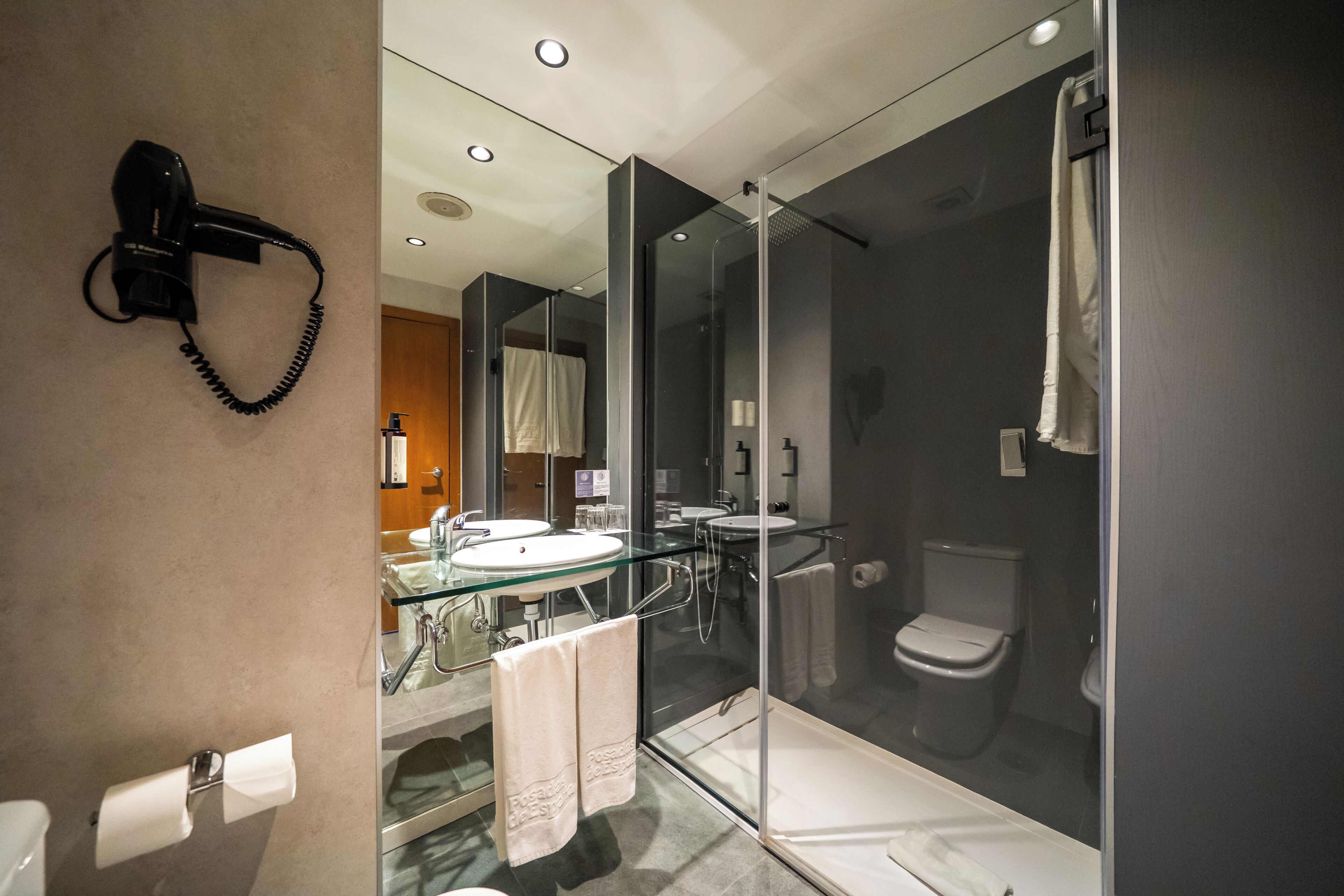 A modern bathroom with a glass shower enclosure, a hairdryer mounted on the wall, and a visible toilet.