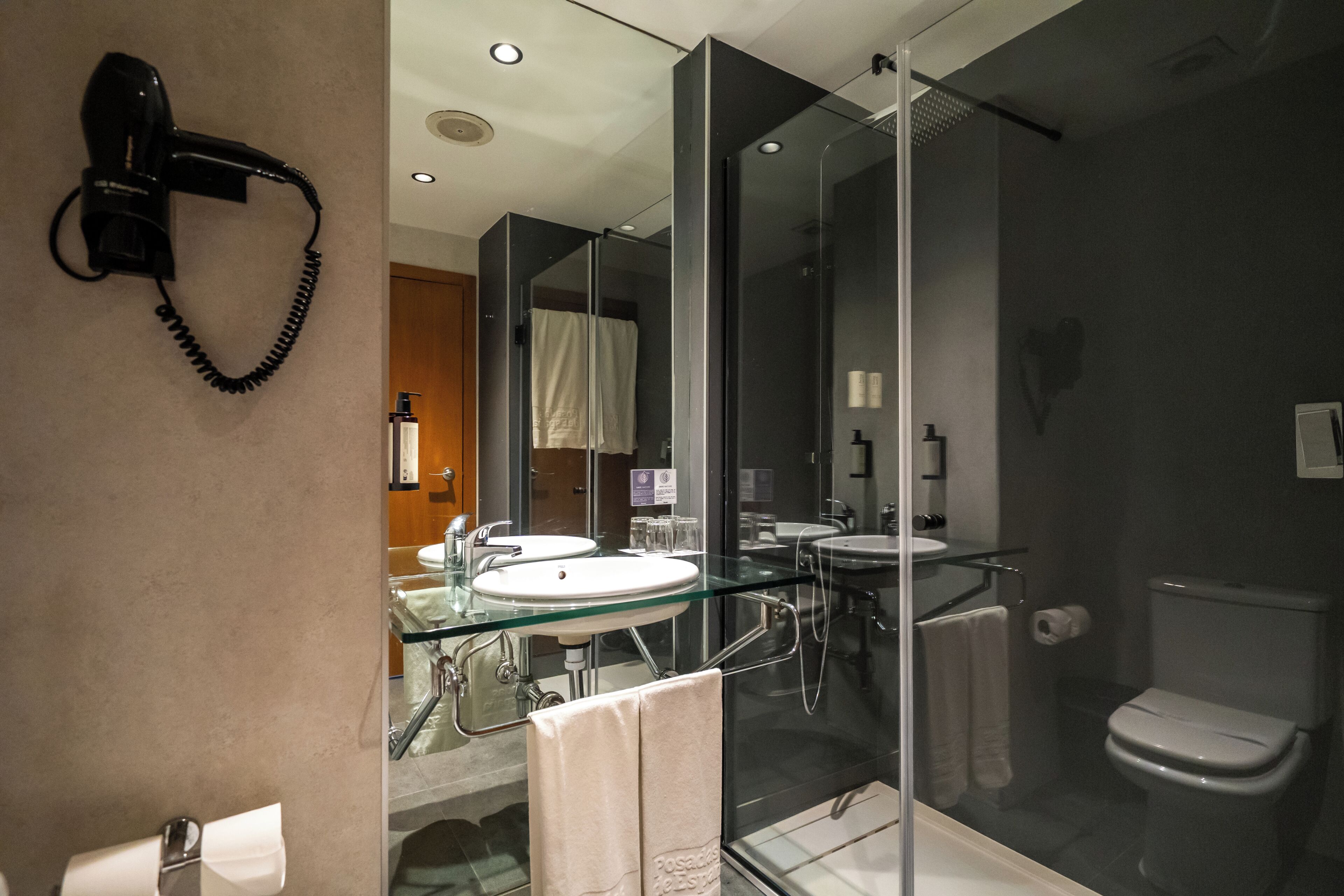 A modern bathroom with a glass-enclosed shower, dual sinks, and a hairdryer mounted on the wall.