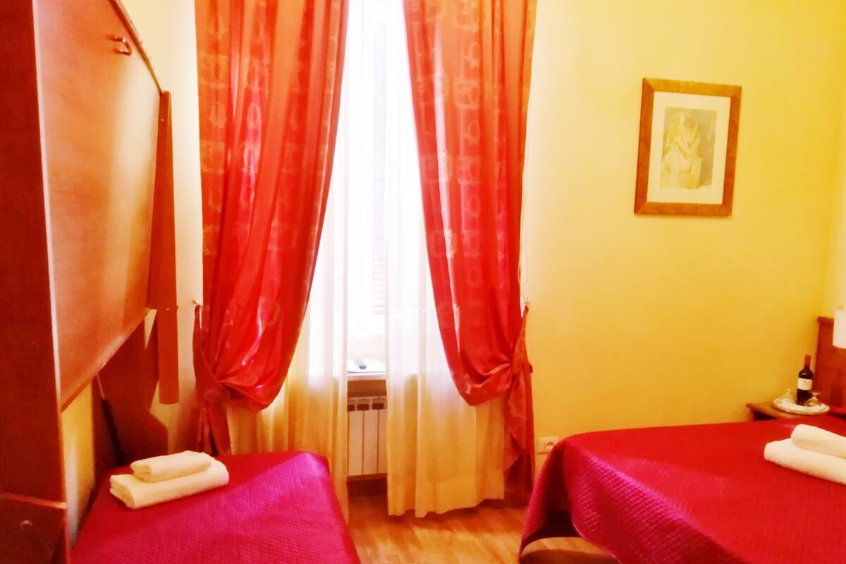 A room with two beds, red curtains, a framed picture on the wall, and a small table with a bottle.