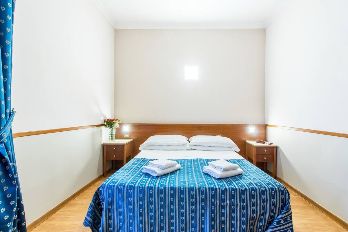 A hotel room with a bed, two bedside tables, a nightstand, and a blue curtain.