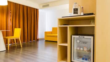 A hotel room with a wooden floor, a yellow chair, a white chair, a wooden shelf with a refrigerator, and a wooden wall.