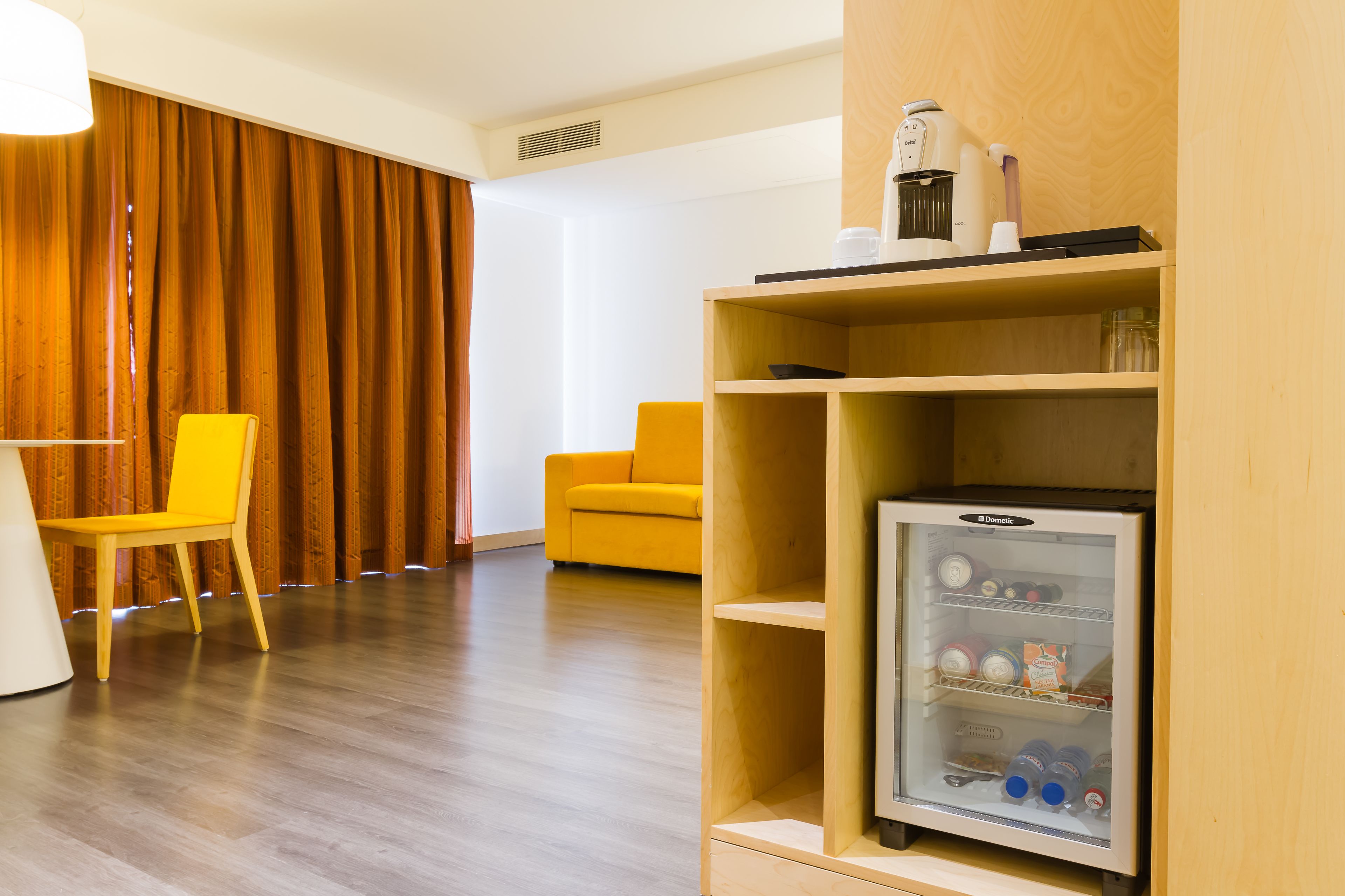 Une chambre d’hôtel avec un plancher en bois, une chaise jaune, une chaise blanche, une étagère en bois avec un réfrigérateur et un mur en bois.