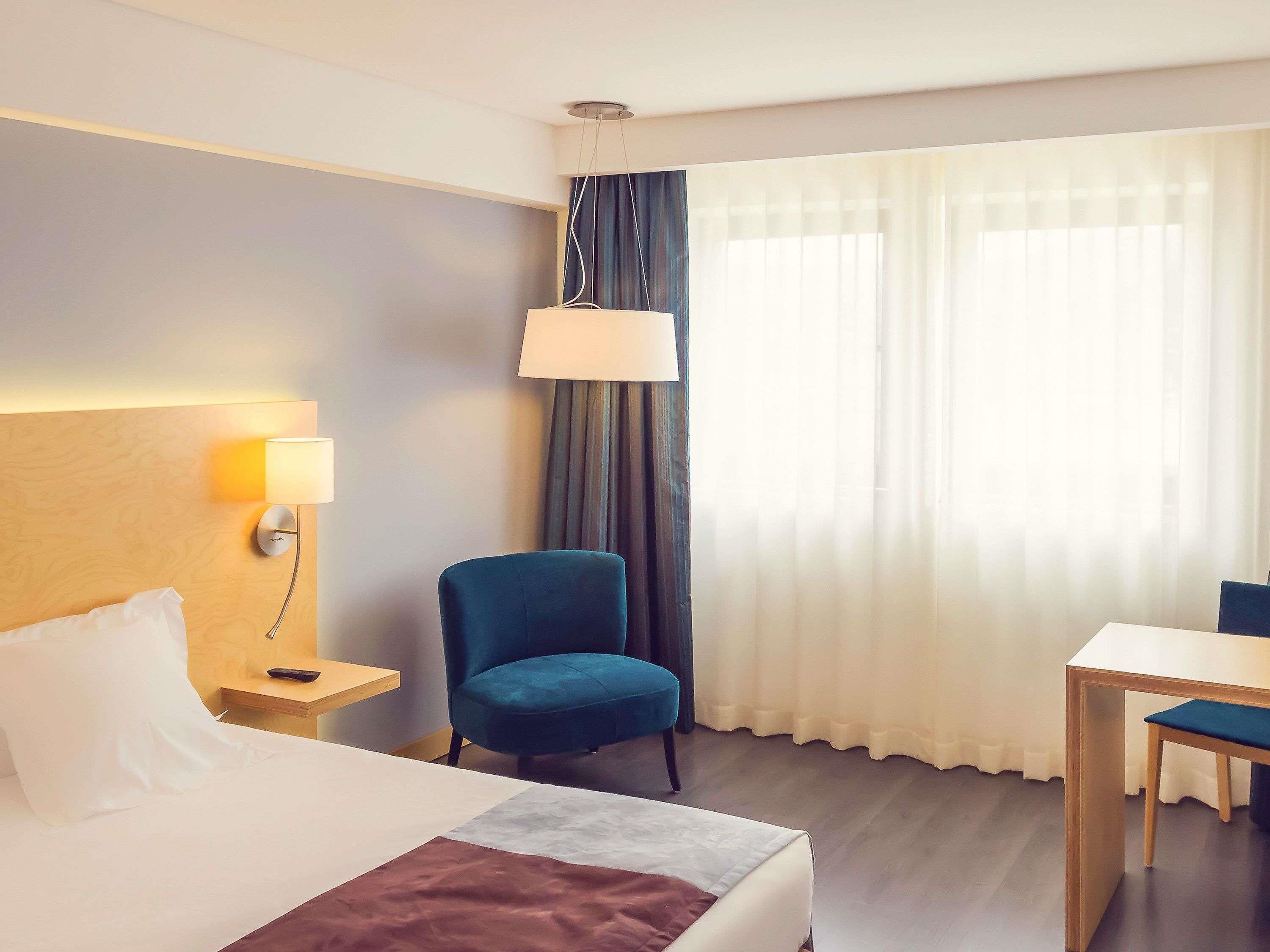 Una habitación de hotel con cama, escritorio, silla, lámpara y ventana con cortinas.
